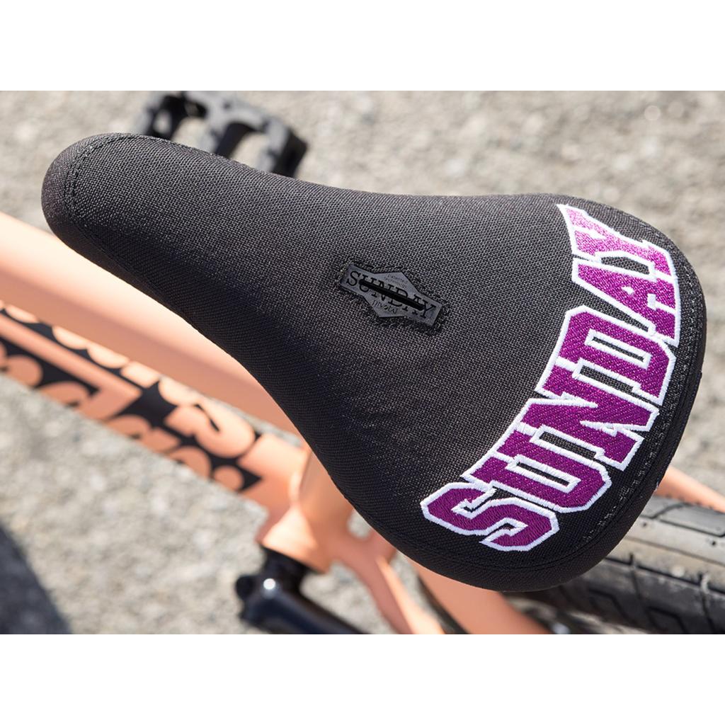 Close-up of a black bicycle seat with "SUNDAY" embroidered in purple and white, mounted on the light peach Sunday Street Sweeper 20 Inch Bike—the Jake Seeley signature chromoly frame.