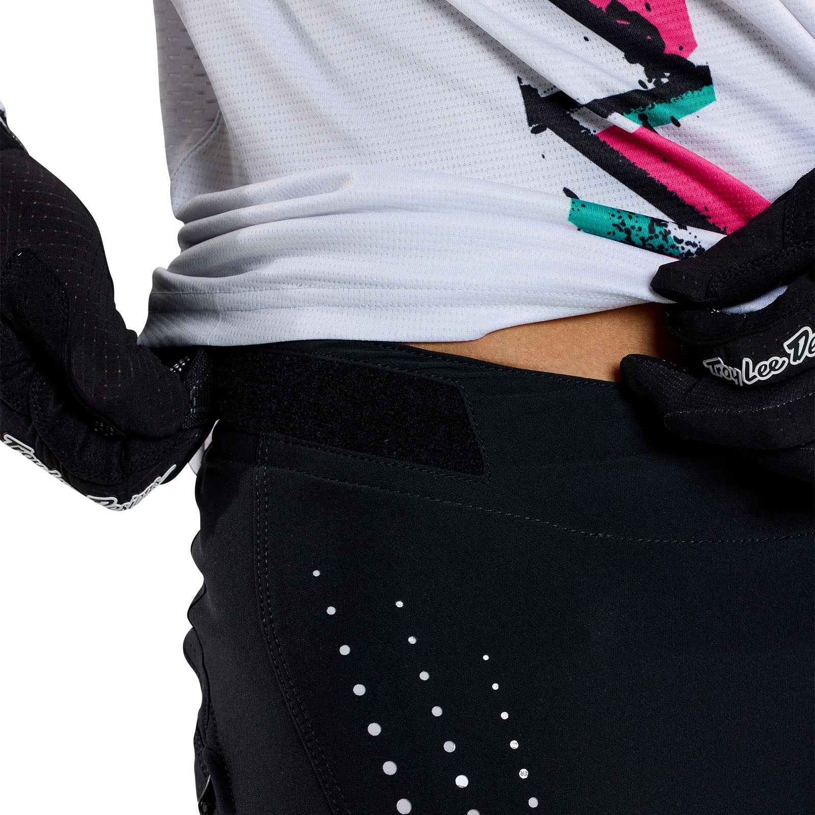 A person adjusts their TLD 25.1 Sprint Pant / Mono Black with gloved hands while wearing a white shirt with an abstract design.