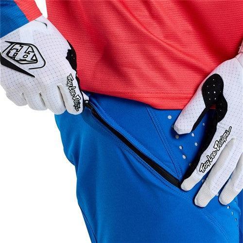 A person in a red and blue outfit with white gloves unzips a pocket on race-ready TLD 25.1 Sprint Pant / Mono Bright Blue, crafted from durable materials.
