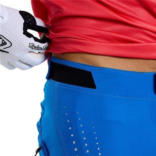A person in a red shirt adjusts the waistband of their TLD 25.1 Sprint Pant in Mono Bright Blue, crafted with durable materials, while wearing a white glove featuring a visible brand logo.