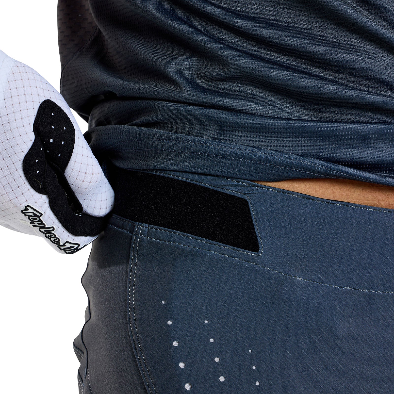 Close-up of someone in a dark sports shirt and TLD 25.1 Sprint Pant / Mono Steel Blue, adjusting a glove labeled "TaylorMade" by their hip with a race-ready black hook and loop fastener.