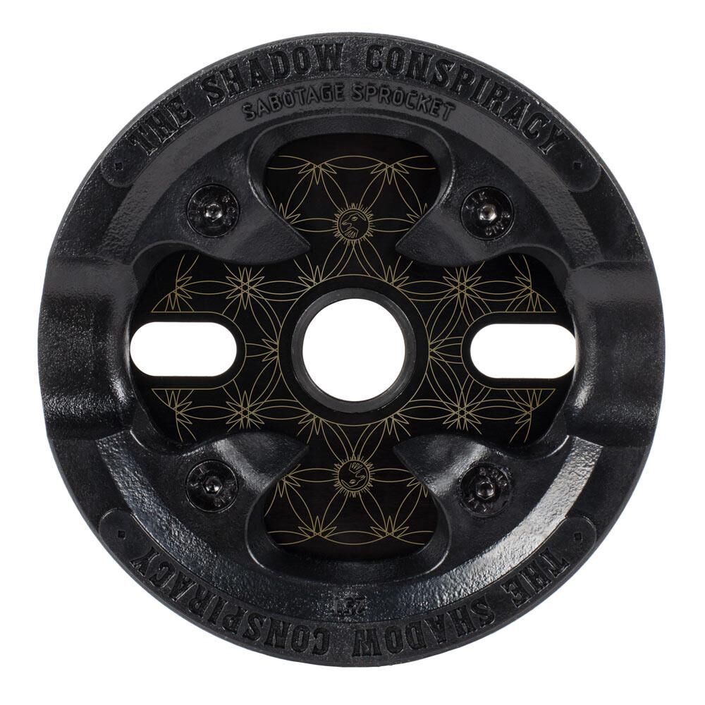 A black metal Shadow Sabotage Maya Edition V2 Sprocket with four bolt holes, a geometric center, and “The Shadow Conspiracy” text around the edge.