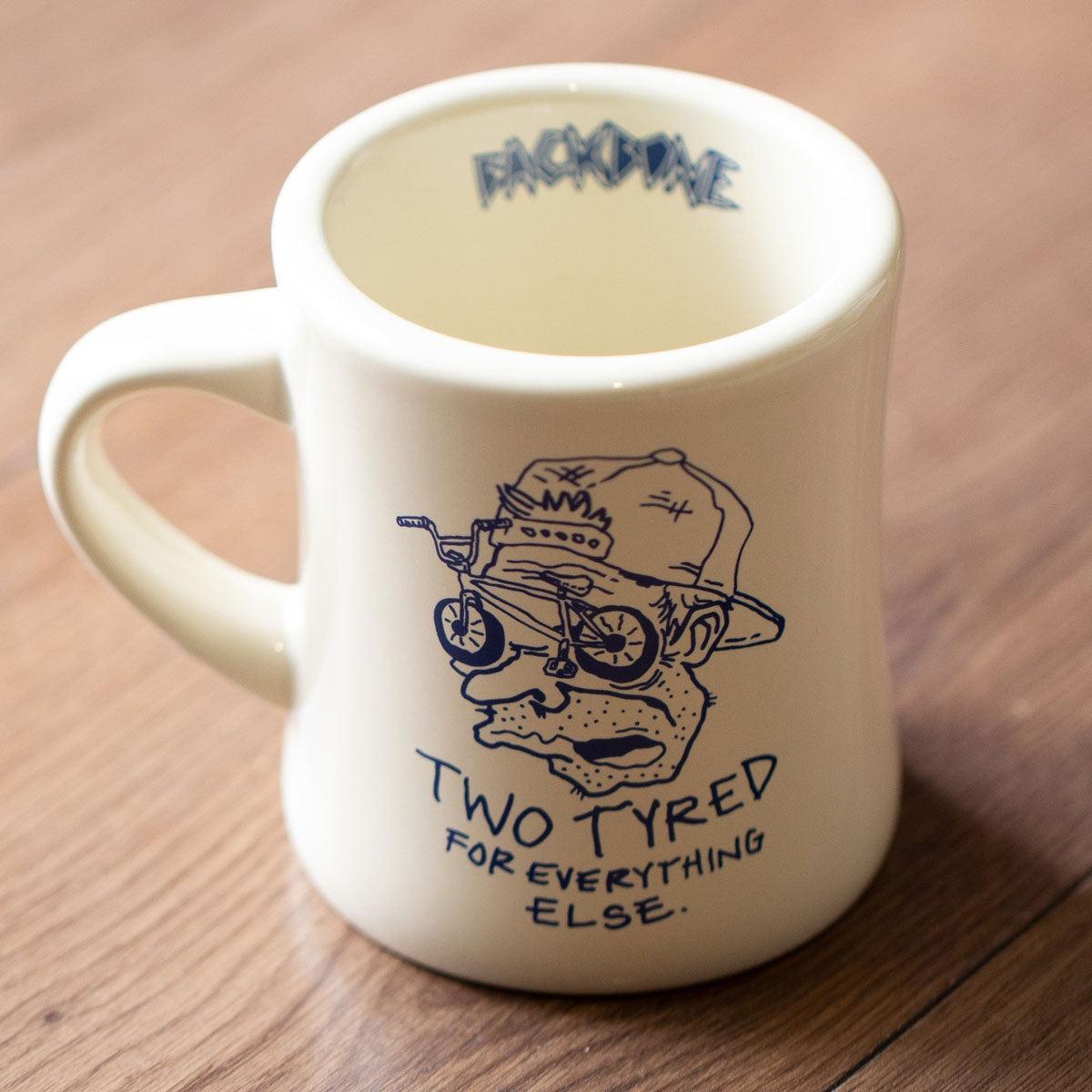 The Backbone Two Tyred Diner Mug is a cream ceramic mug featuring a cartoon face with bicycle glasses and "TWO TYRED FOR EVERYTHING ELSE." A quirky BMX gift for bikers, it's perfect for your coffee break.