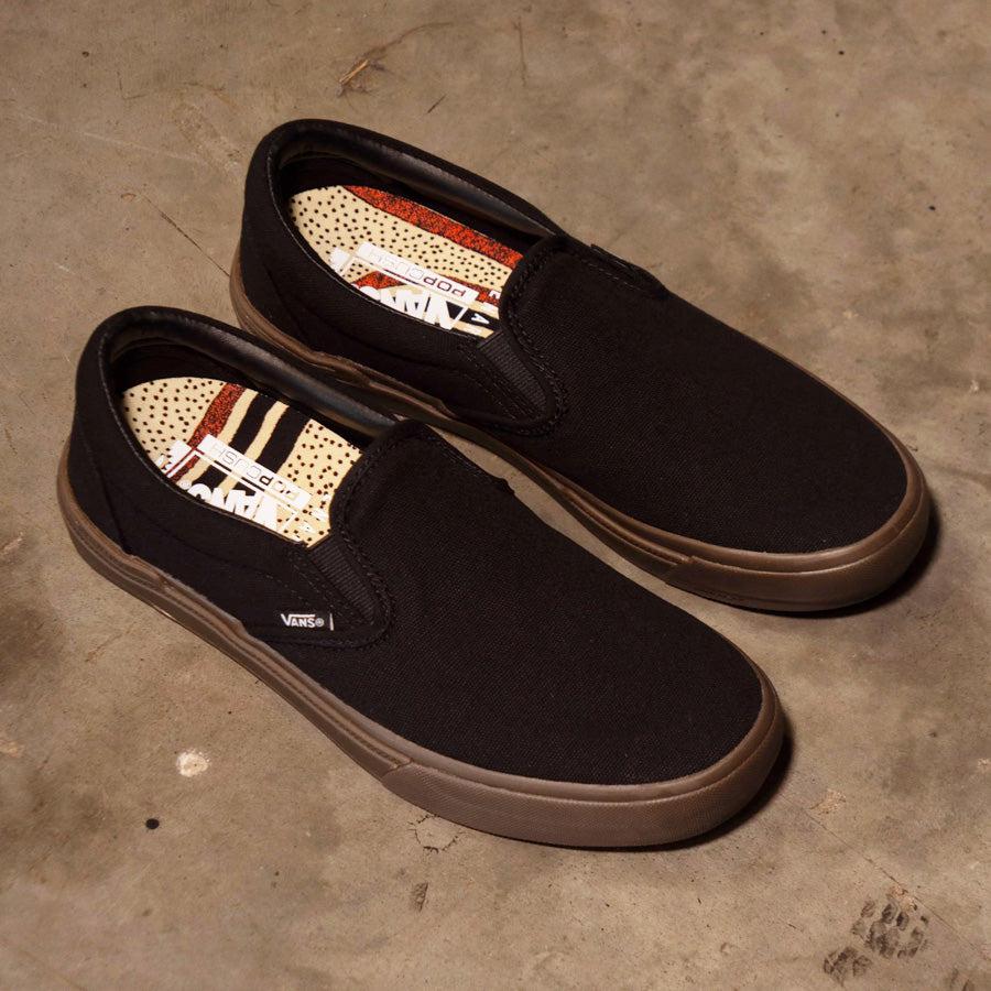 A pair of black slip-on Vans BMX Slip On Shoes (Dennis Enarson) Black/Multi on a concrete floor, emphasizing durability.