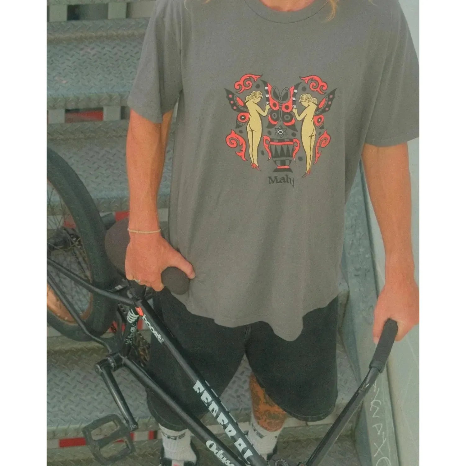 A person stands on a metal staircase in Cape Town, holding a black BMX bike and wearing the Mahd Wesley Tee with a vibrant traditional tattoo-inspired graphic, paired with black shorts.