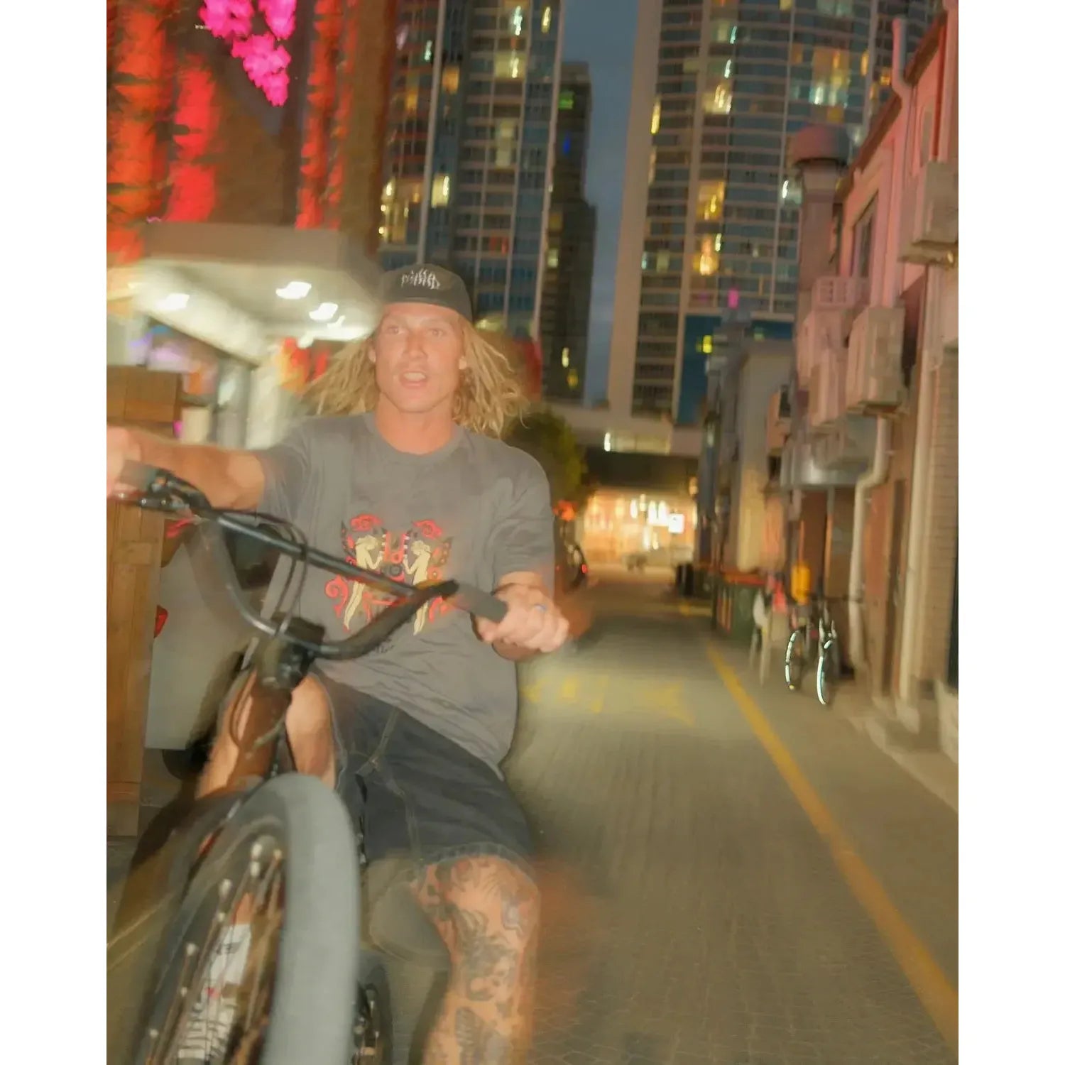 Wearing the Mahd Wesley Tee, a person with long blond hair and traditional tattoos rides a bike on one wheel down a Cape Town street at dusk, tall buildings rising in the background.