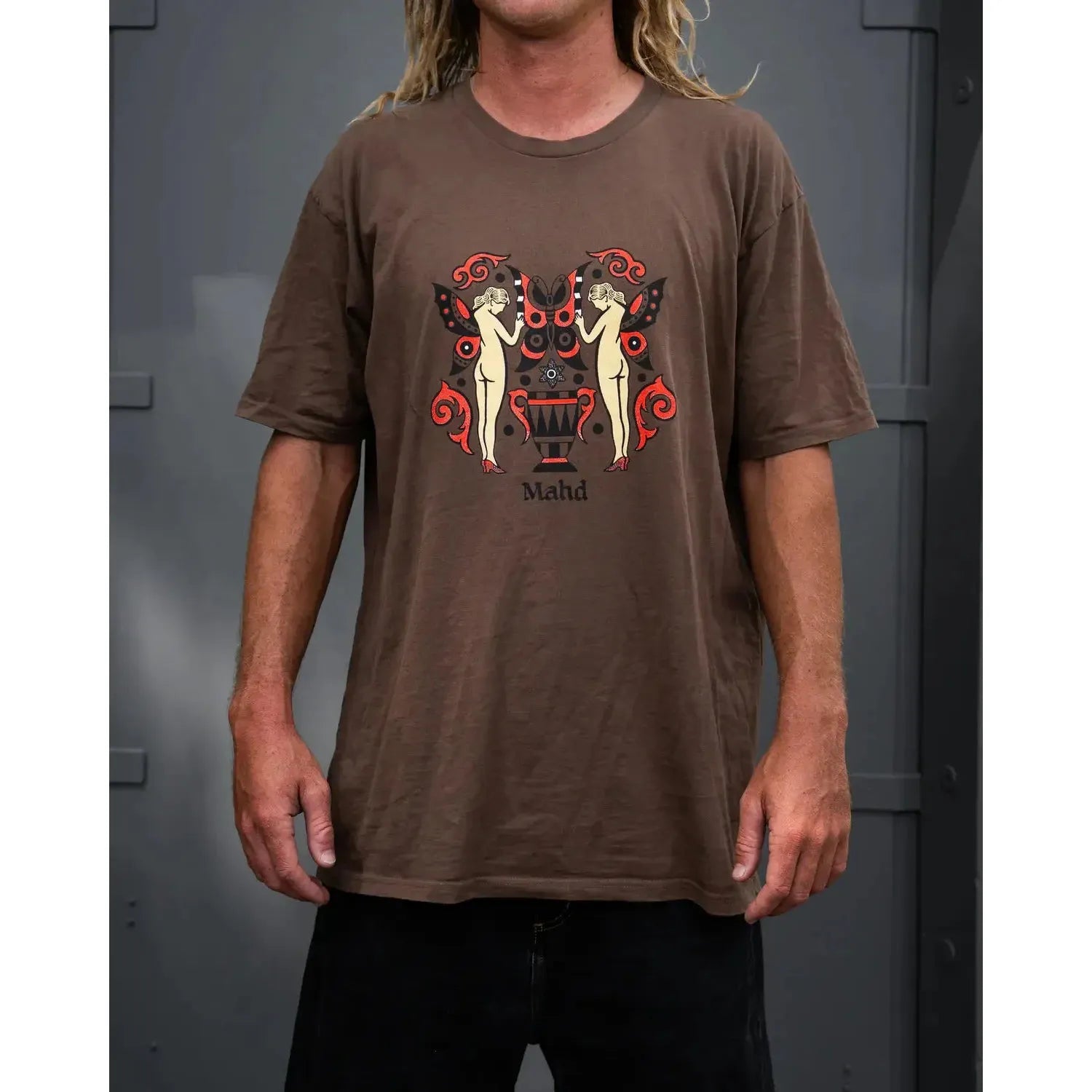 Someone wears the Mahd Wesley Tee, a brown shirt featuring an artistic graphic of two nude figures and a decorative urn in traditional tattoo style, with "Mahd" below the design.