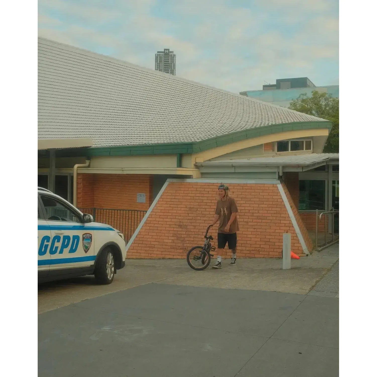 In Cape Town, a BMX rider stands by a brick building near a GCPD police car, wearing the Mahd Wesley Tee featuring traditional tattoo style graphics.