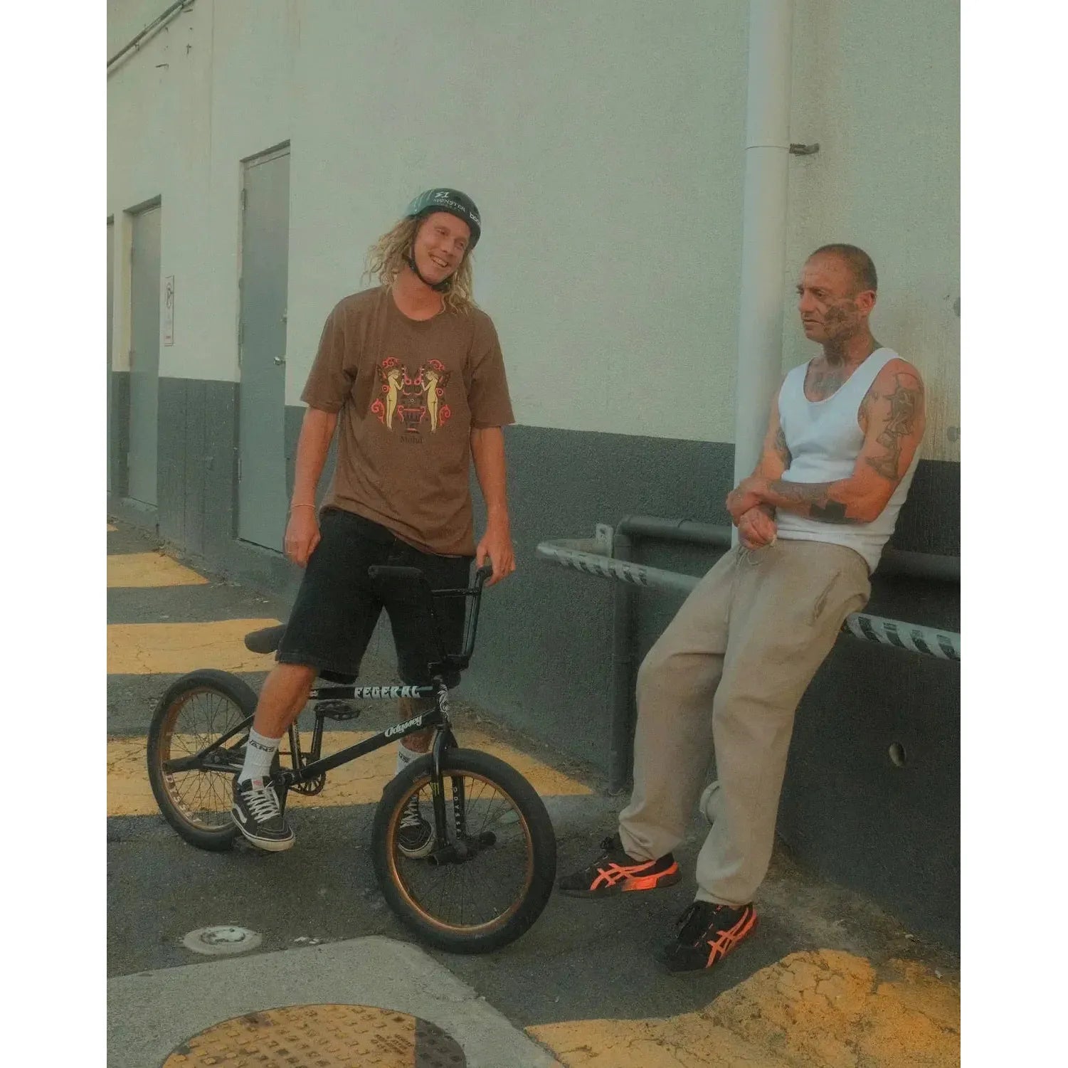Two men outside a building in Cape Town: one stands with a BMX bike, wearing a helmet and casual clothes featuring the Mahd Wesley Tee; the other leans against a railing in a tank top and sweatpants.