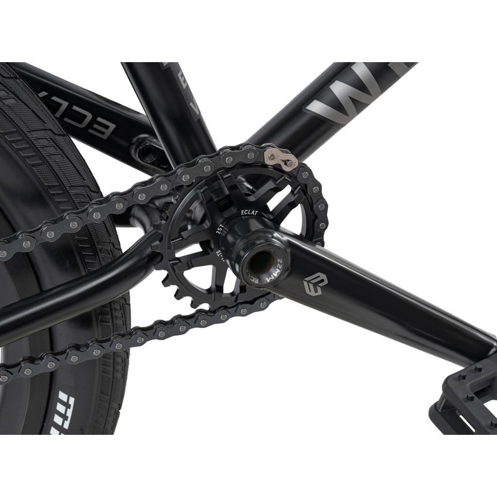 Close-up of the Wethepeople 20 inch Envy Carbonic LTD Bike’s crankset and chain, highlighting the pedal arm, chainring, and rear tire. The black components display brand markings for a sleek appearance.