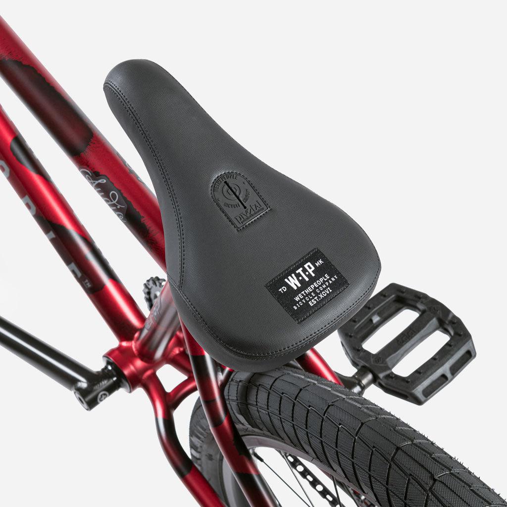 Close-up of the black bicycle seat with brand logos and text, mounted on the red chromoly frame of the Wethepeople Audio 22 Inch Bike, showing part of the rear wheel and pedal of this trail bike.