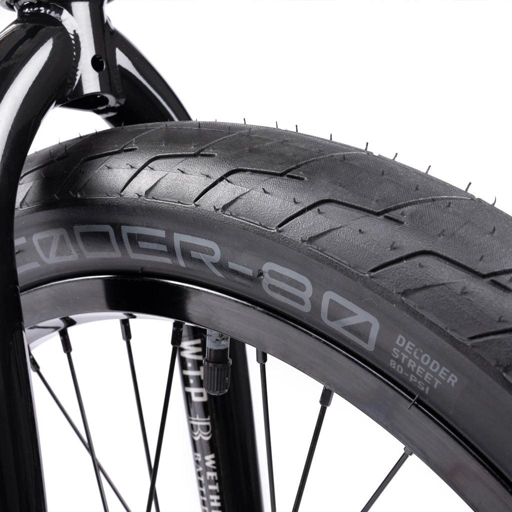 Close-up of the Wethepeople Battleship 20 Inch Bike's black BMX tire mounted on a rim, showing its tread pattern and "DECODER-80 DECODER STREET 80 PSI" sidewall text. Eclat components provide durable, high-performance riding.