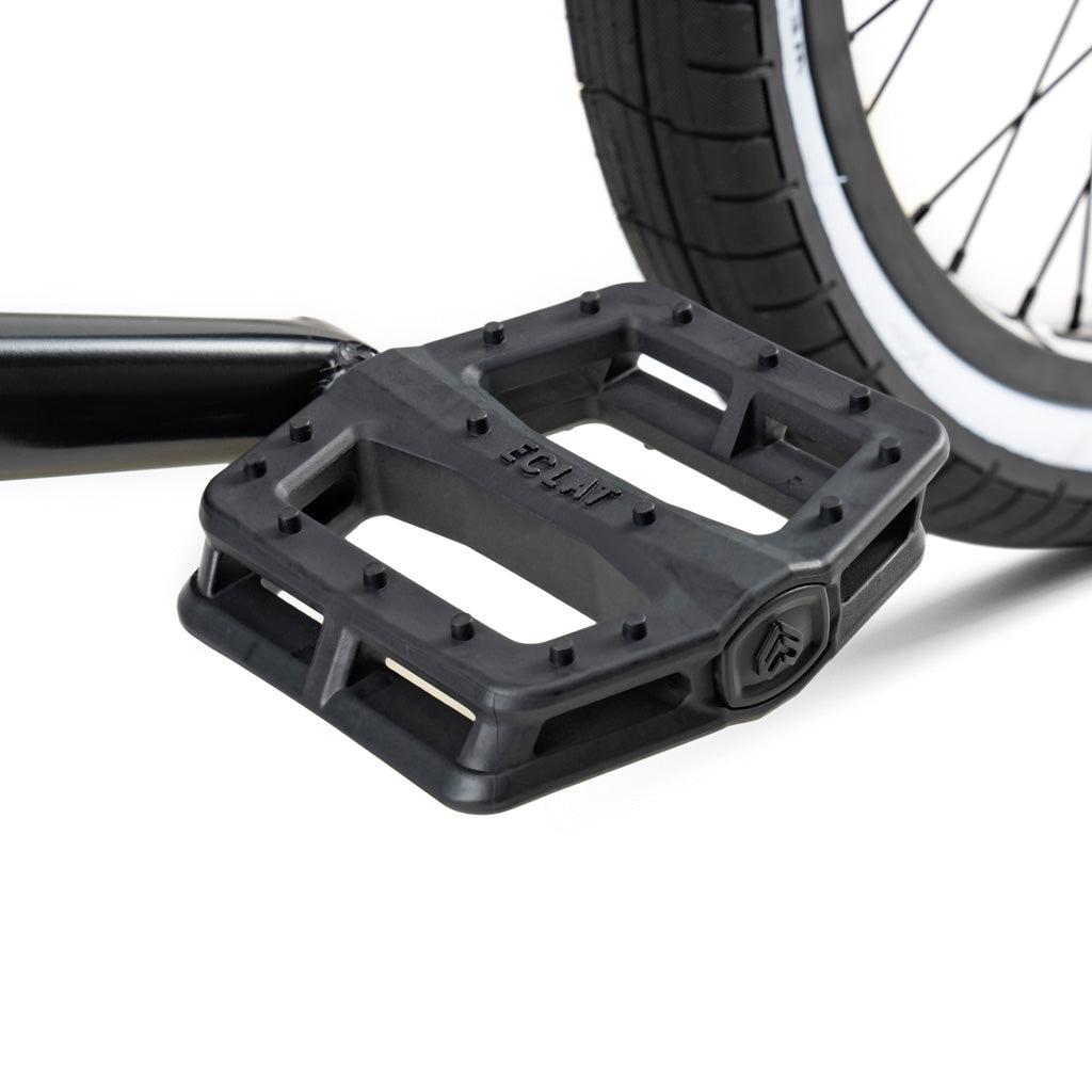 Close-up of a black plastic pedal attached to the crank of a Wethepeople Battleship 20 Inch Bike, shown next to part of its BMX wheel with a black tire and white stripe.