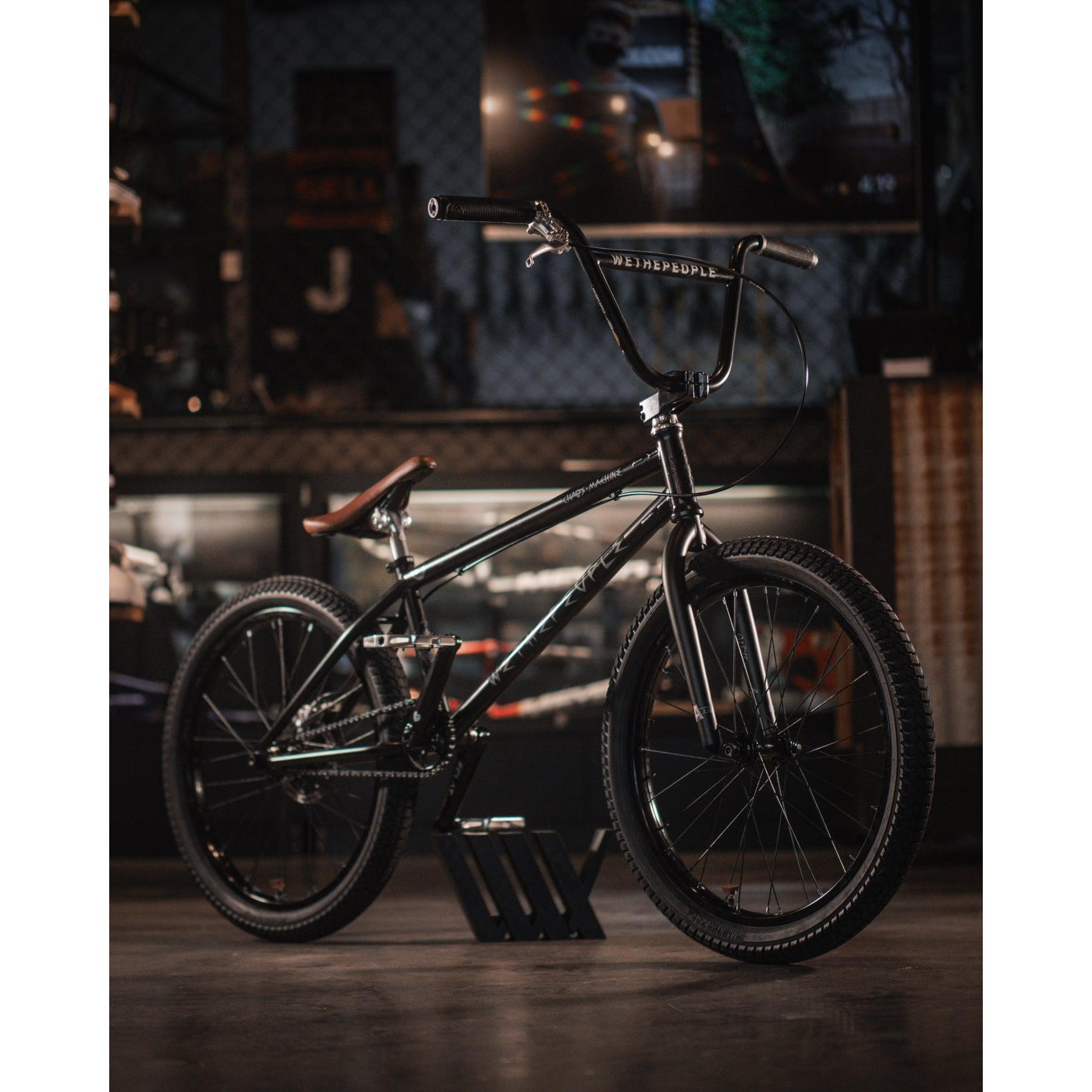 A black BMX bike with "WeThePeople" branding, also known as the Wethepeople Chaos Machine 22 Inch Custom Bike, is placed on a stand in a dimly lit indoor setting. The bike features thick tires, a brown leather saddle, and a clean chain.