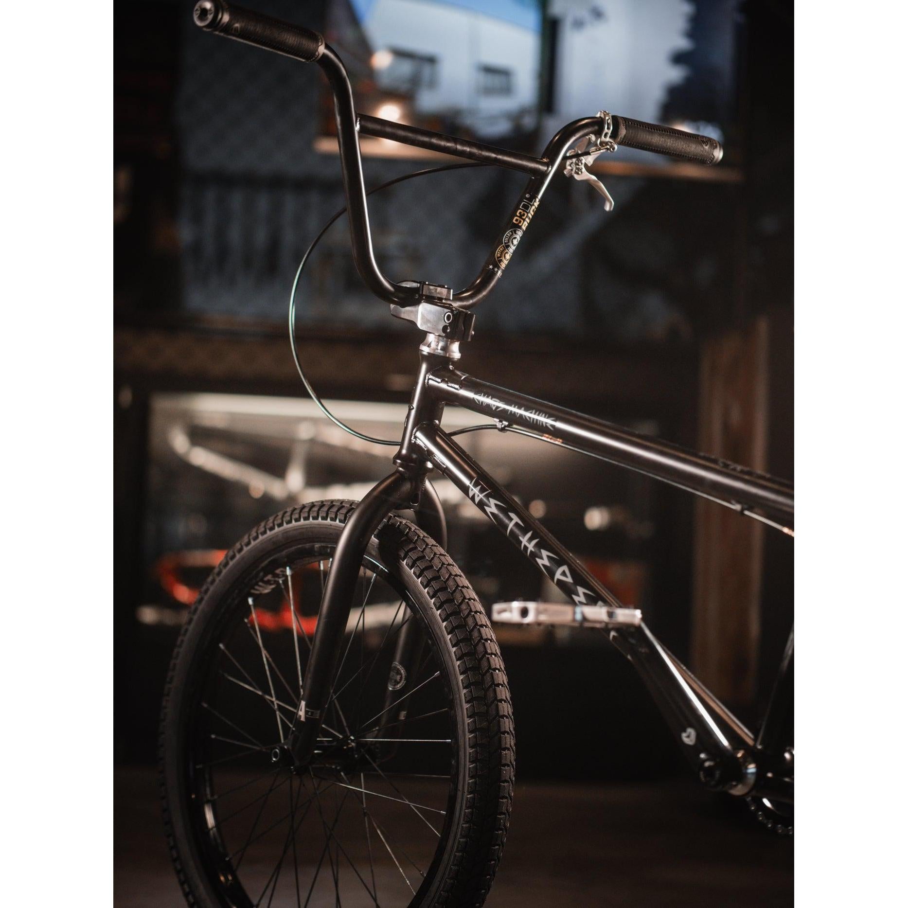 A close-up of a BMX bike in a dimly lit environment, showcasing the handlebars and front tire, highlighted by the sleek design of the Wethepeople Chaos Machine 22 Inch Custom Bike.