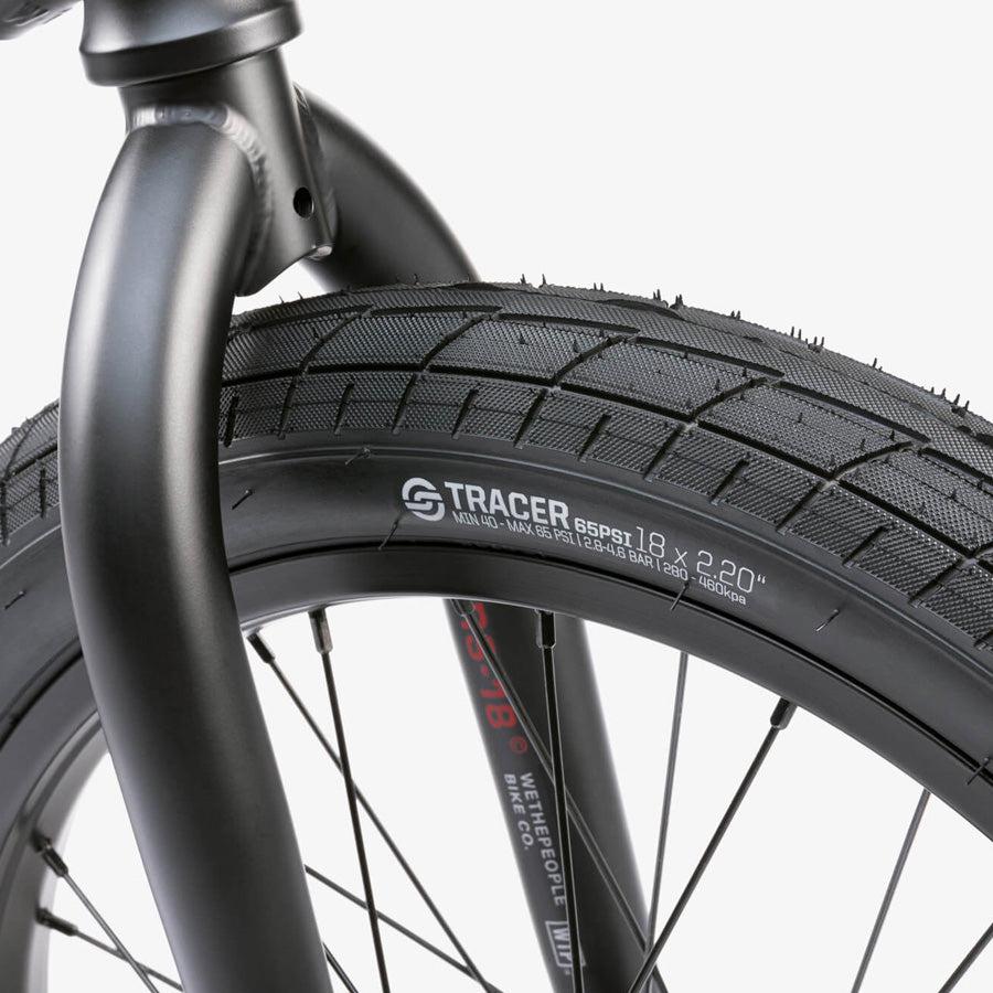 Close-up of the Wethepeople CRS 18 Inch Bike front wheel with a black Tracer 20 x 2.20 tire (tread pattern and valve stem visible) on a black rim with spokes—ideal for an 18 inch BMX ride.