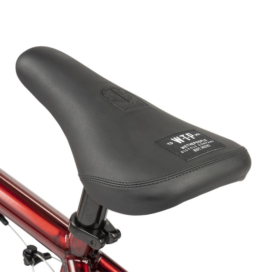 Close-up of a black bicycle seat with stitched logo and label, mounted on the metallic red frame of the Wethepeople CRS 18 Inch Bike.