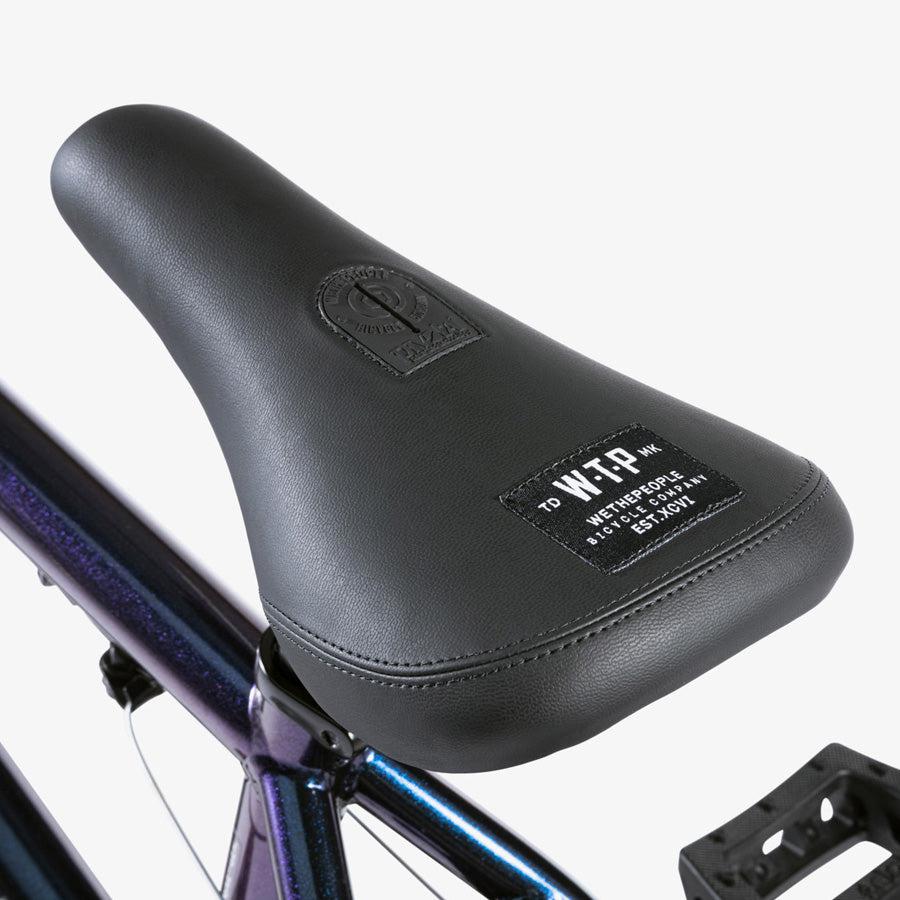 A close-up of a black bicycle seat with branding, mounted on the metallic frame of a Wethepeople CRS 18 Inch Bike, against a white background.