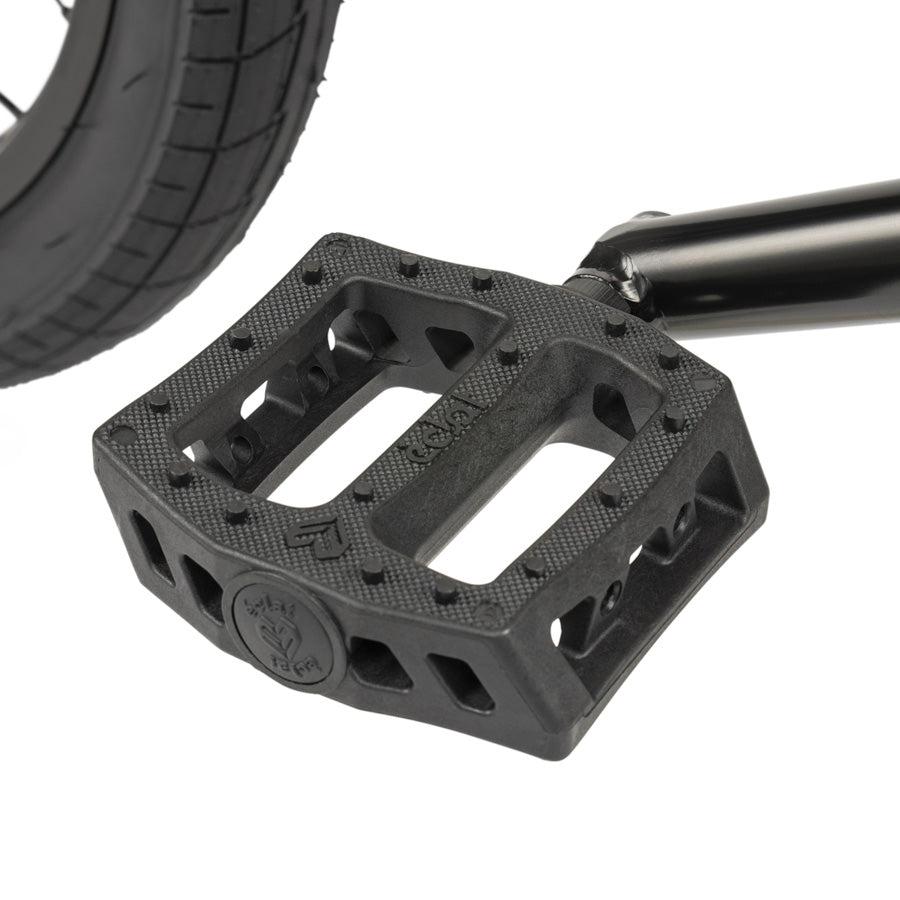 Close-up of a black plastic bicycle pedal on a crank arm, with a bike tire behind—showing perfect detail for the Wethepeople CRS 18 Inch Bike, an ideal choice for kids’ BMX riding.