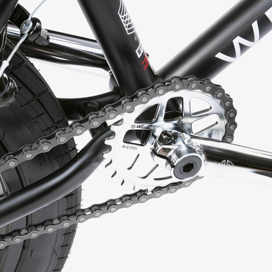 Close-up of the crankset, chainring, and chain on a black Wethepeople CRS 18 Inch Bike frame, with the rear wheel and tire in view—showcasing details perfect for an 18 inch BMX.