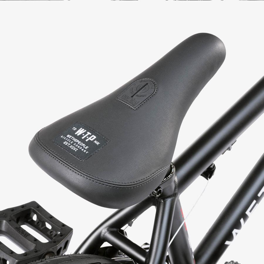 Close-up of a black bicycle seat with white branding, attached to a black Wethepeople CRS 18 Inch Bike frame. The plain white background highlights this stylish kids' BMX bike.