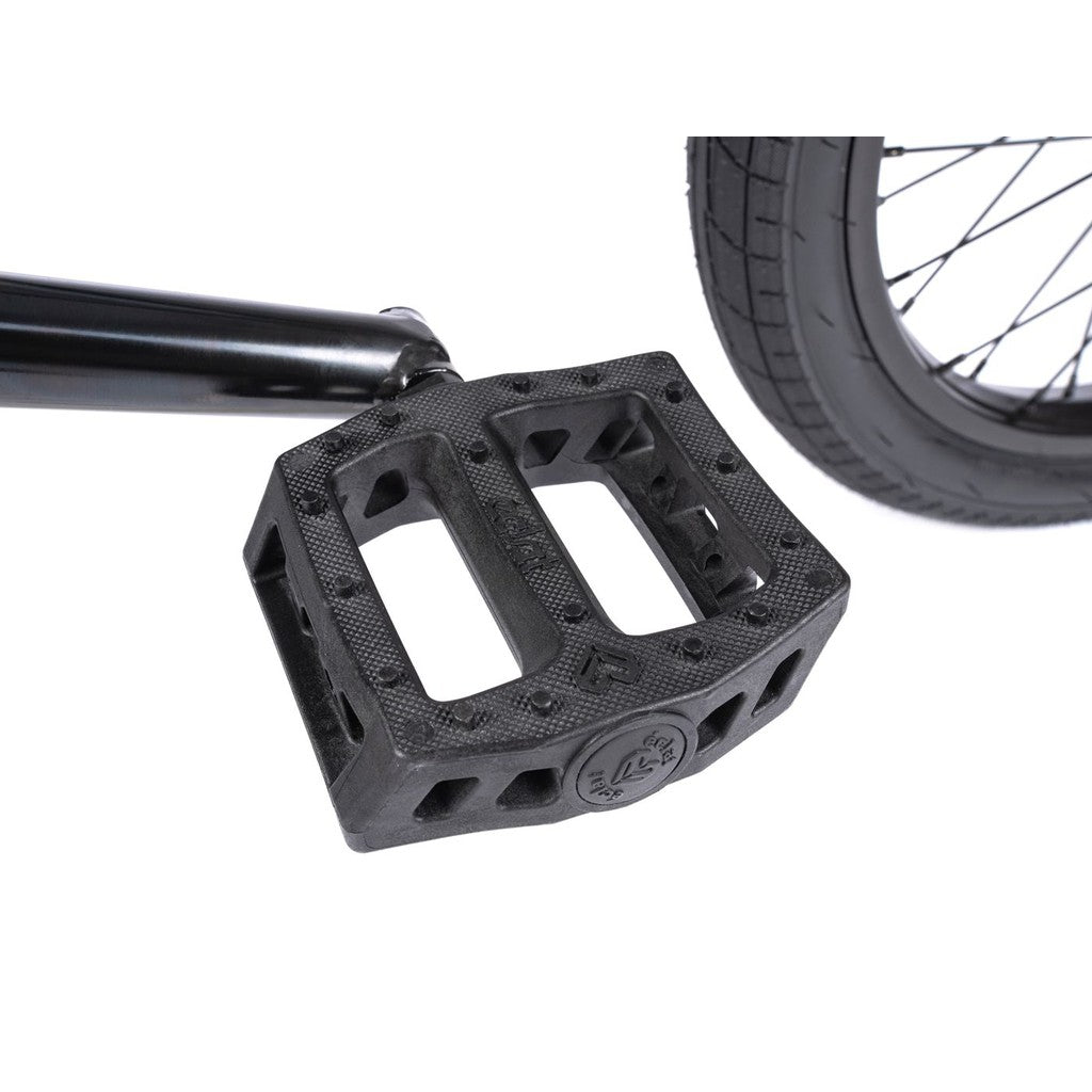Close-up of a black plastic bicycle pedal attached to a crank arm on the Wethepeople CRS FS 18 Inch Bike, with part of the rear wheel and tire visible in the background.