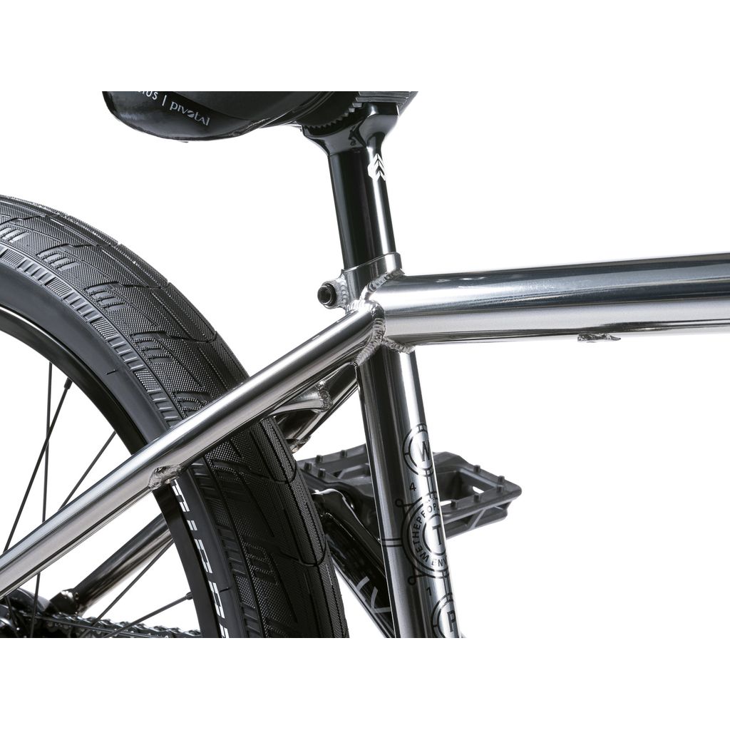 Close-up of a silver frame featuring the rear wheel, seat post, and part of the seat. The Wethepeople Envy 20 Inch Bike shines with a metallic finish and treaded black tire, embodying BMX mastery.