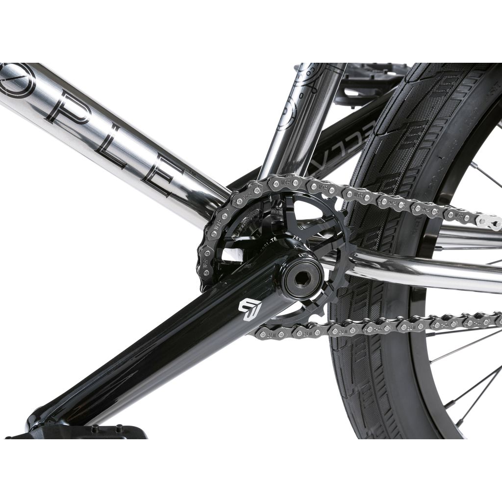 Close-up of the Wethepeople Envy 20 Inch Bike's drivetrain, featuring a black crankset, silver chain, and rear tire. The metallic frame with engraved logos enhances the sleek elegance of Eclat Tibia Cranks for high-performance cycling.