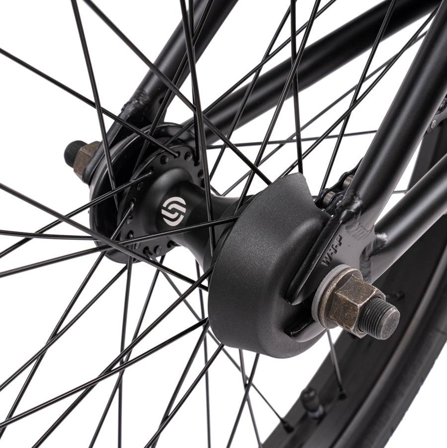 Close-up of the Wethepeople Justice 20 Inch Bike's rear wheel hub and spokes, showing the axle, nuts, and a device attached to the frame. The black and metallic components are pictured against a plain white background.