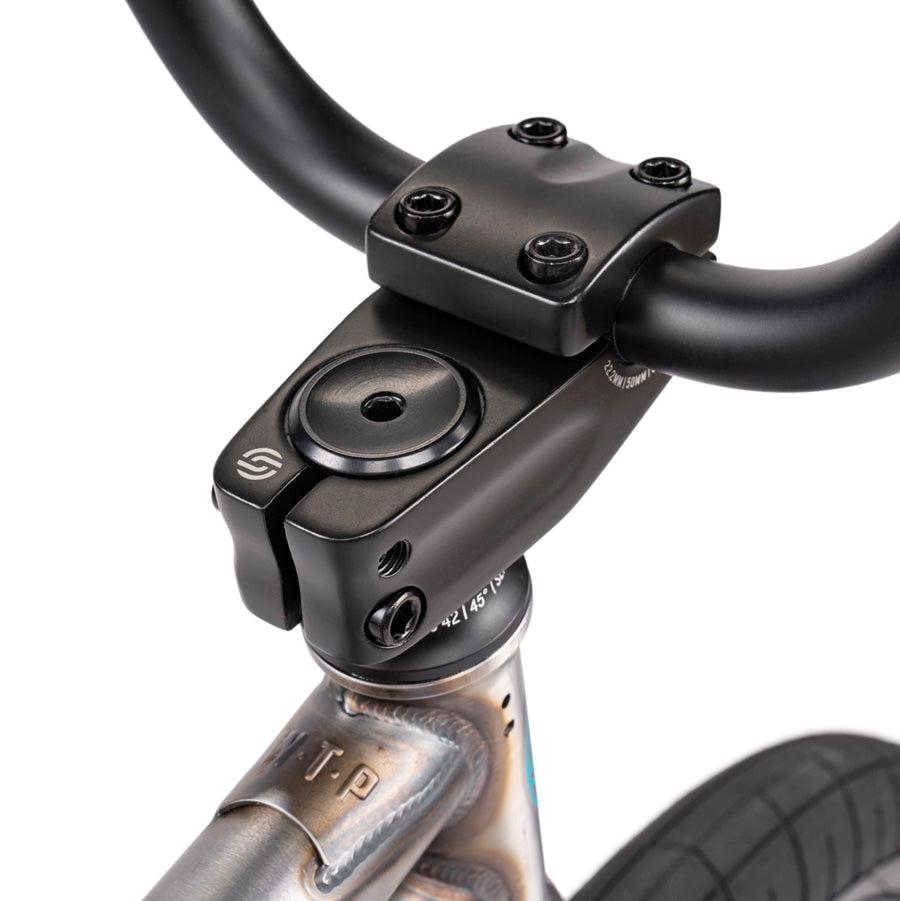 Close-up view of a black bicycle stem connecting the handlebars to the steerer tube on the Wethepeople Justice 20 Inch Bike. The image showcases intricate details, including bolts and branding on both the handlebar and stem—hallmarks of top-of-the-line BMX components. A tire is partially visible.