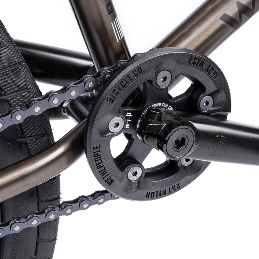 Close-up of the Wethepeople Justice 20 Inch Bike drivetrain system, showing a black chain ring, pedal arm, and part of the tire. The chain is visible and the frame features a dark metallic finish, reminiscent of an Urban BMX Bike with top-of-the-line BMX components.