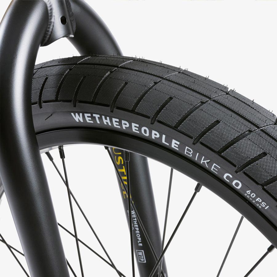 Close-up of a Wethepeople Justice 20 Inch Bike wheel showcasing a black tire labeled "WETHEPEOPLE BIKE CO" mounted on an elegant black metal frame featuring premium BMX components.