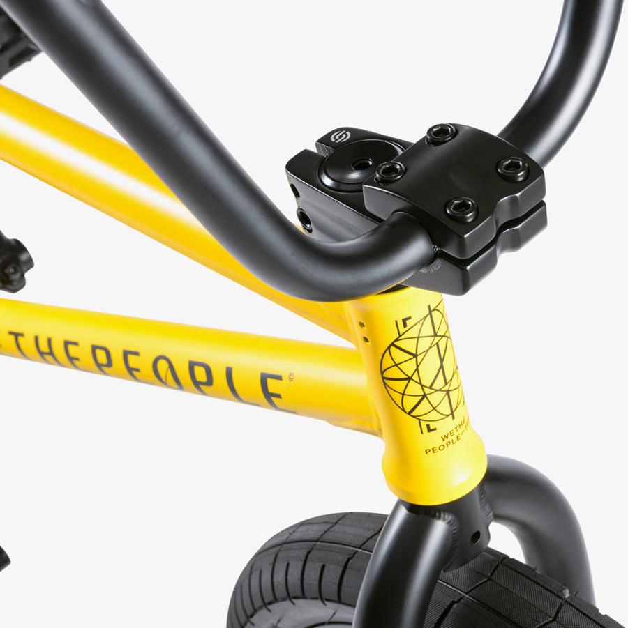 Close-up of a yellow urban BMX bike frame with black handlebars and a tire. The frame features the text "THE PEOPL" alongside a geometric design, representing the premium BMX components you'd expect from the Wethepeople Justice 20 Inch Bike.