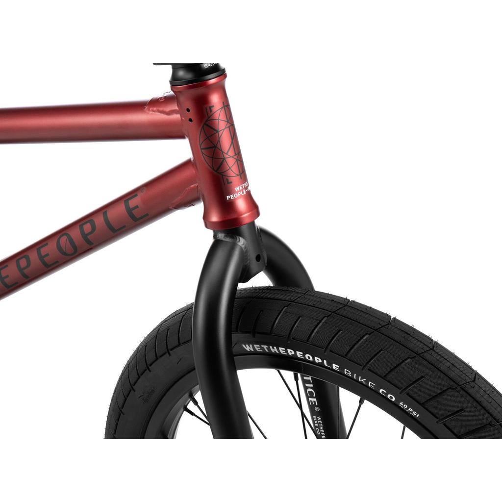 Close-up of the Wethepeople Justice 20 Inch Bike's red frame and black front fork; its front tire displays "WETHEPEOPLE BIKE CO" branding on the sidewall.