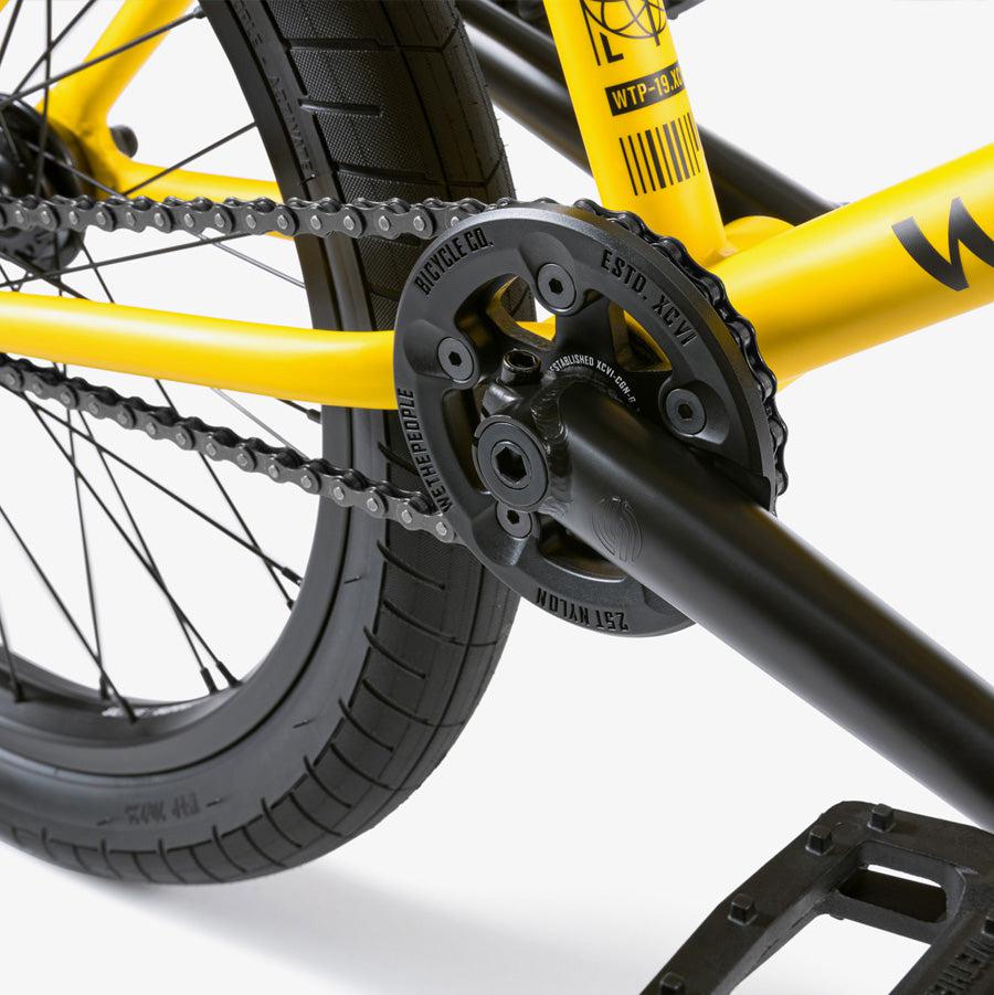 Close-up view of a yellow Wethepeople Justice 20 Inch Bike's chains and gears, featuring top-of-the-line BMX components including the rear wheel and pedal crank mechanism.