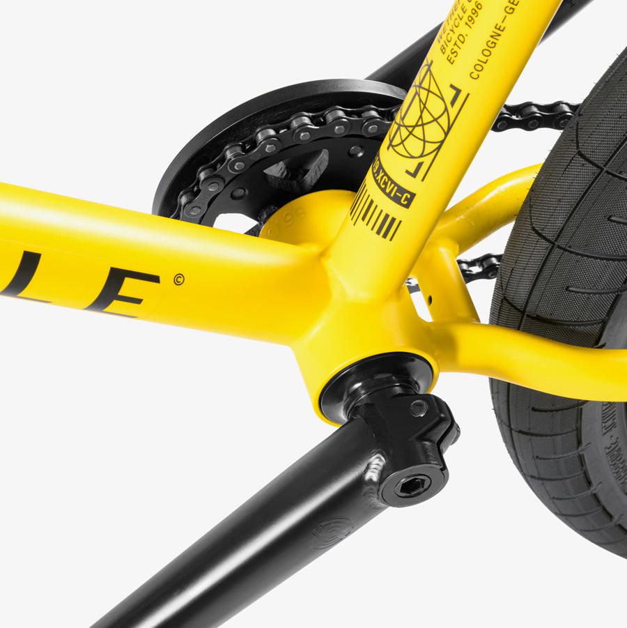 Close-up of a yellow Wethepeople Justice 20 Inch Bike's bottom bracket and crankset, showcasing the chainring and black crank arm, with part of the rear tire visible.