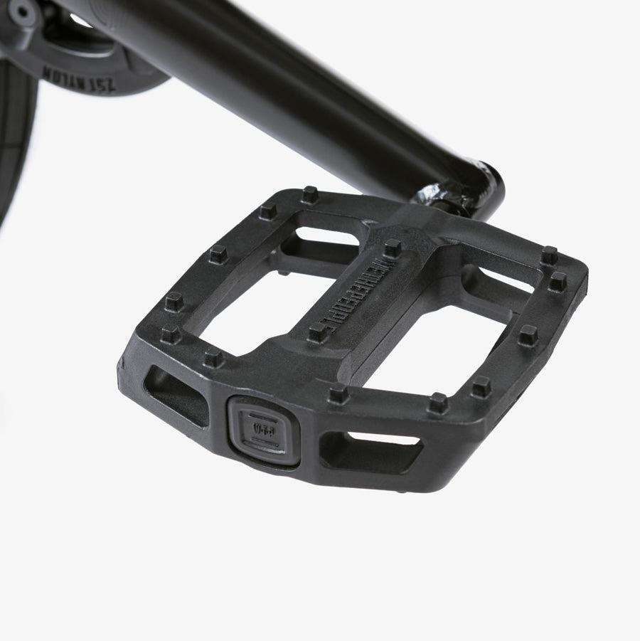 Close-up of a black bicycle pedal attached to a crank arm on the Wethepeople Justice 20 Inch Bike. The pedal features a wide, flat surface with small protrusions for grip.