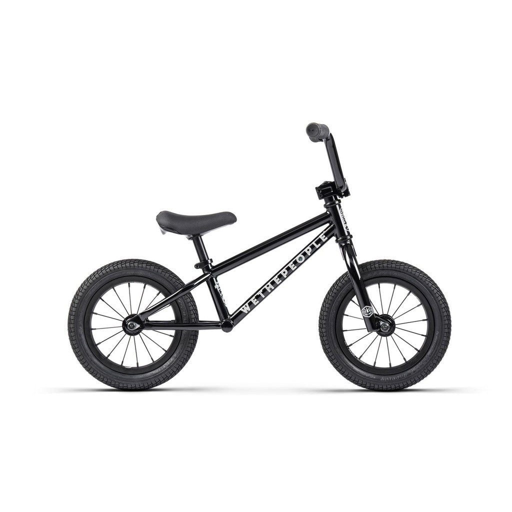 The Wethepeople Prime 12 Inch Balance Bike is a small black BMX-style balance bike with no pedals, featuring a padded seat, thick tires, and "WETHEPEOPLE" branding on the frame.