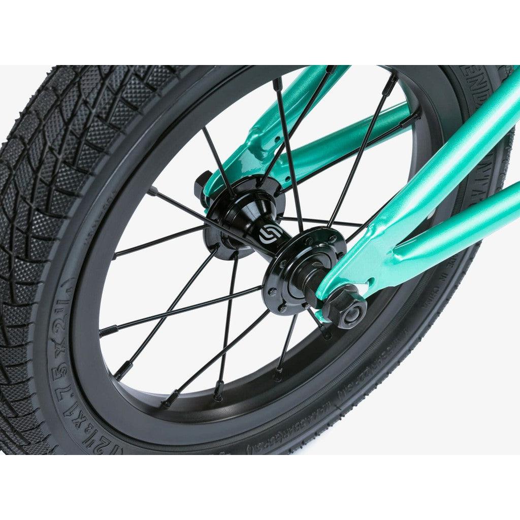 Close-up of the Wethepeople Prime 12 Inch Balance Bike wheel featuring black spokes, a black hub, teal fork, and a textured black tire with visible tread pattern.
