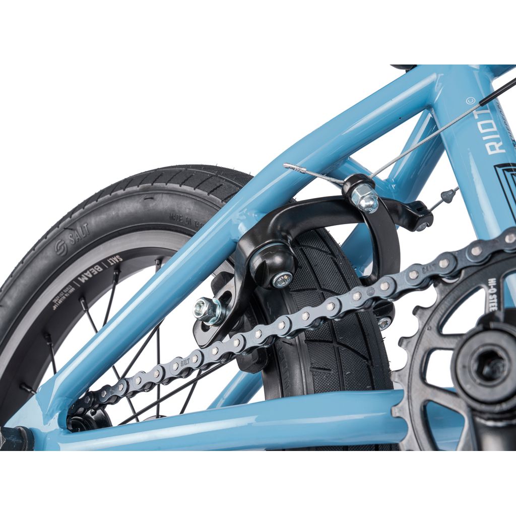 Close-up view of a Wethepeople Riot 14 Inch Bike's rear wheel, featuring the blue tire, brake mechanism, chain, and part of the chromoly cranks.