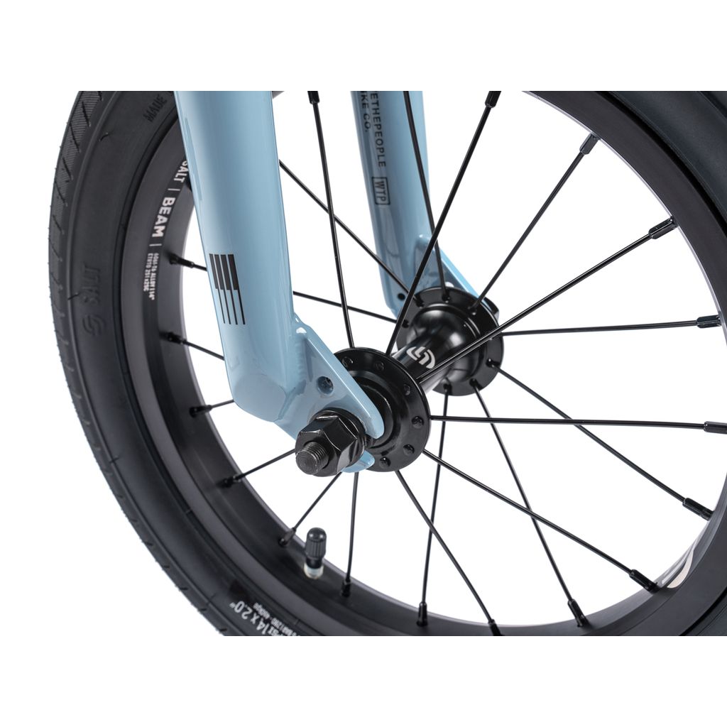 Close-up view of the Wethepeople Riot 14 Inch Bike's front wheel showing tire, spokes, axle bolt, fork, a light blue frame, black components, and durable Chromoly cranks.