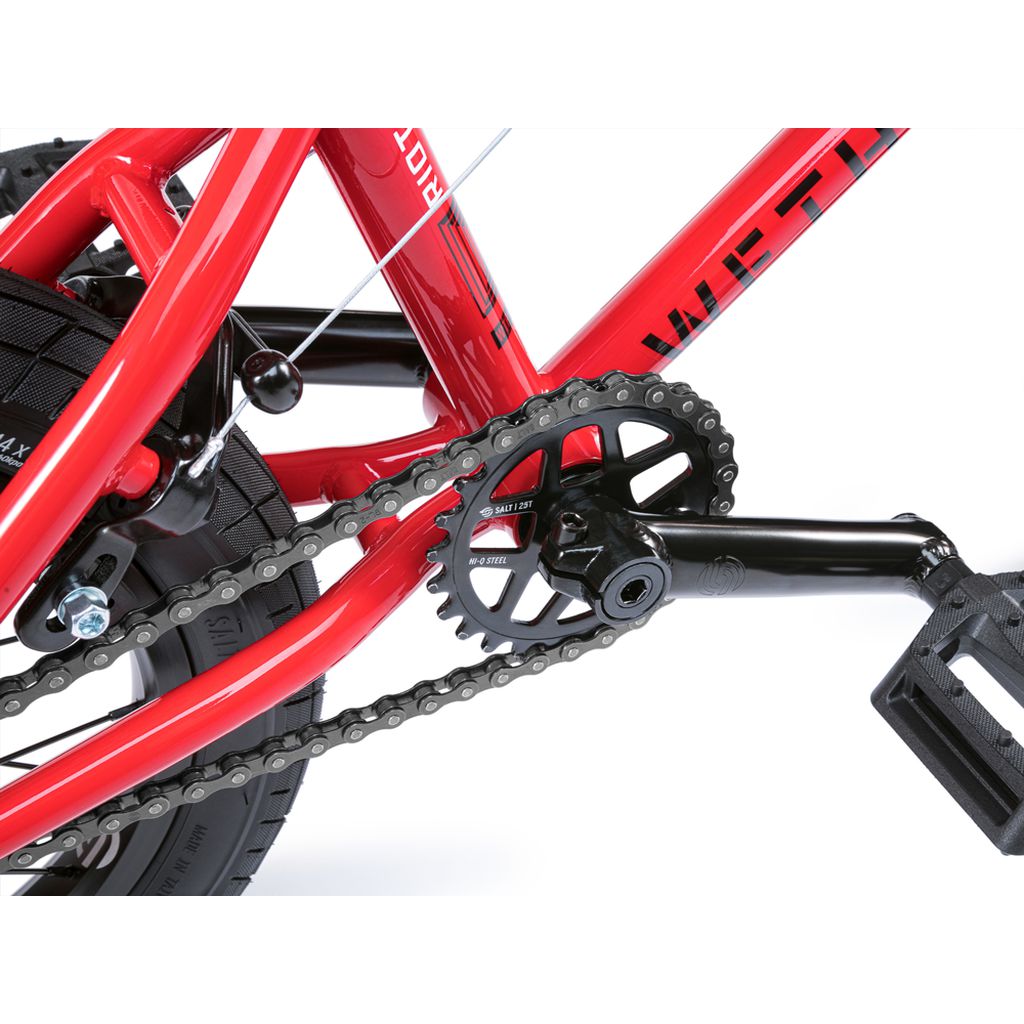Close-up of the Wethepeople Riot 14 Inch Bike's red chain, black Chromoly cranks, and pedal, highlighting the gears and part of the rear wheel.