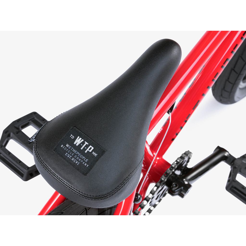 A close-up of the black seat with label, mounted on the red frame of the Wethepeople Riot 14 Inch Bike near the pedals, chain, and robust Chromoly cranks.