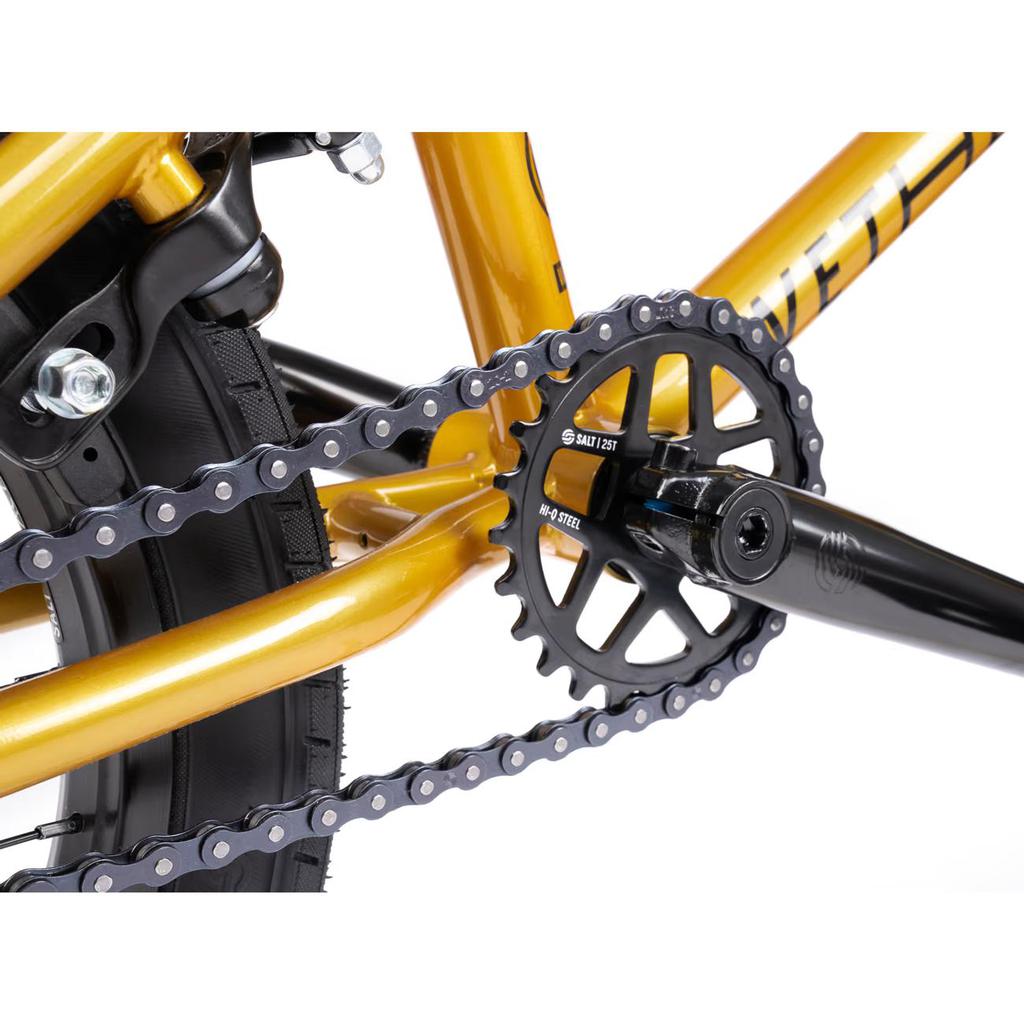 Close-up of a gold-painted bike frame featuring black Salt Rookie Tubular Chromoly Cranks, matching chainring and chain. A large tire tread is visible in the background, ideal for BMX enthusiasts or proud owners of a Wethepeople Seed 16 Inch Bike.