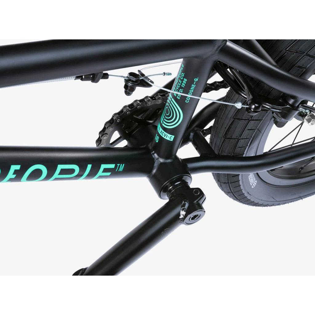 Close-up of a Wethepeople Seed 16 Inch Bike black frame highlighting the bottom bracket, pedal cranks, tire, and a segment of a teal decal.