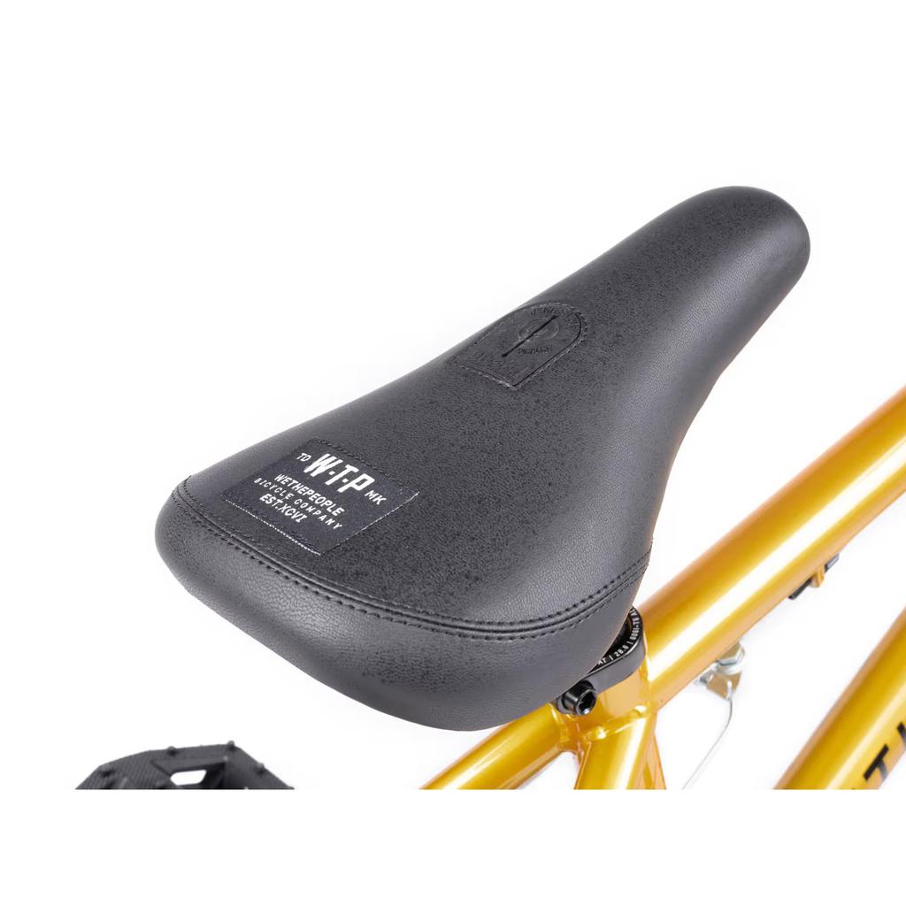Close-up of a black seat with a fabric label on the Wethepeople Seed 16 Inch Bike, featuring a metallic gold frame, ideal for BMX shredders.