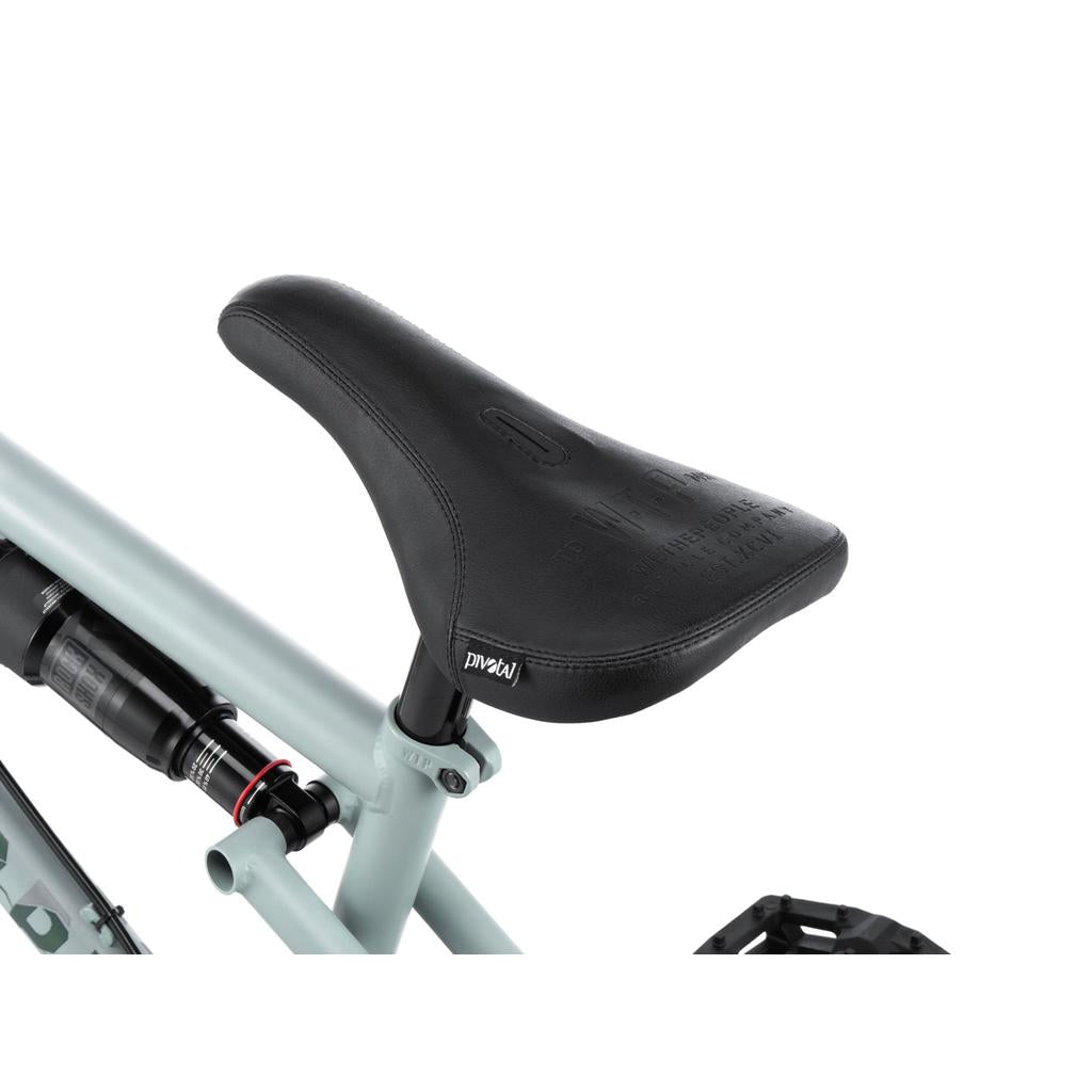 Close-up of the Wethepeople Swampmaster 20 Inch Bike shows a black saddle on a light gray frame with BMX seat post, pedal, and rear suspension partly visible.