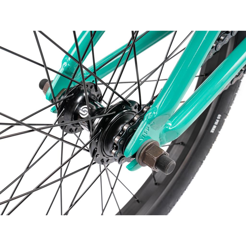 Close-up of a Wethepeople Thrillseeker 20 Inch Bike’s rear wheel hub, with black spokes and a turquoise frame, focusing on the axle and part of the tire.