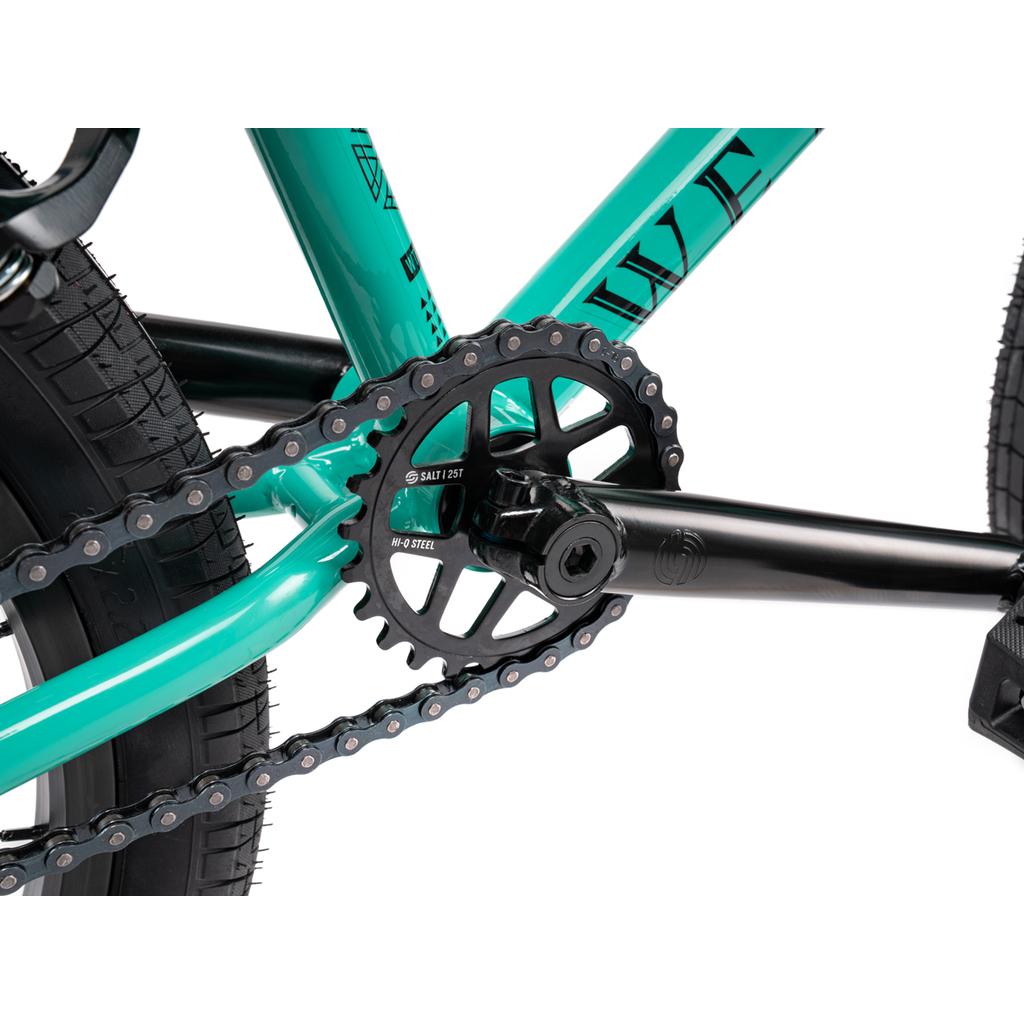 Close-up of the Wethepeople Thrillseeker 20 Inch Bike’s teal Hi-Ten frame, black crank arm, chainring, and chain, highlighting its detailed BMX components and connections.
