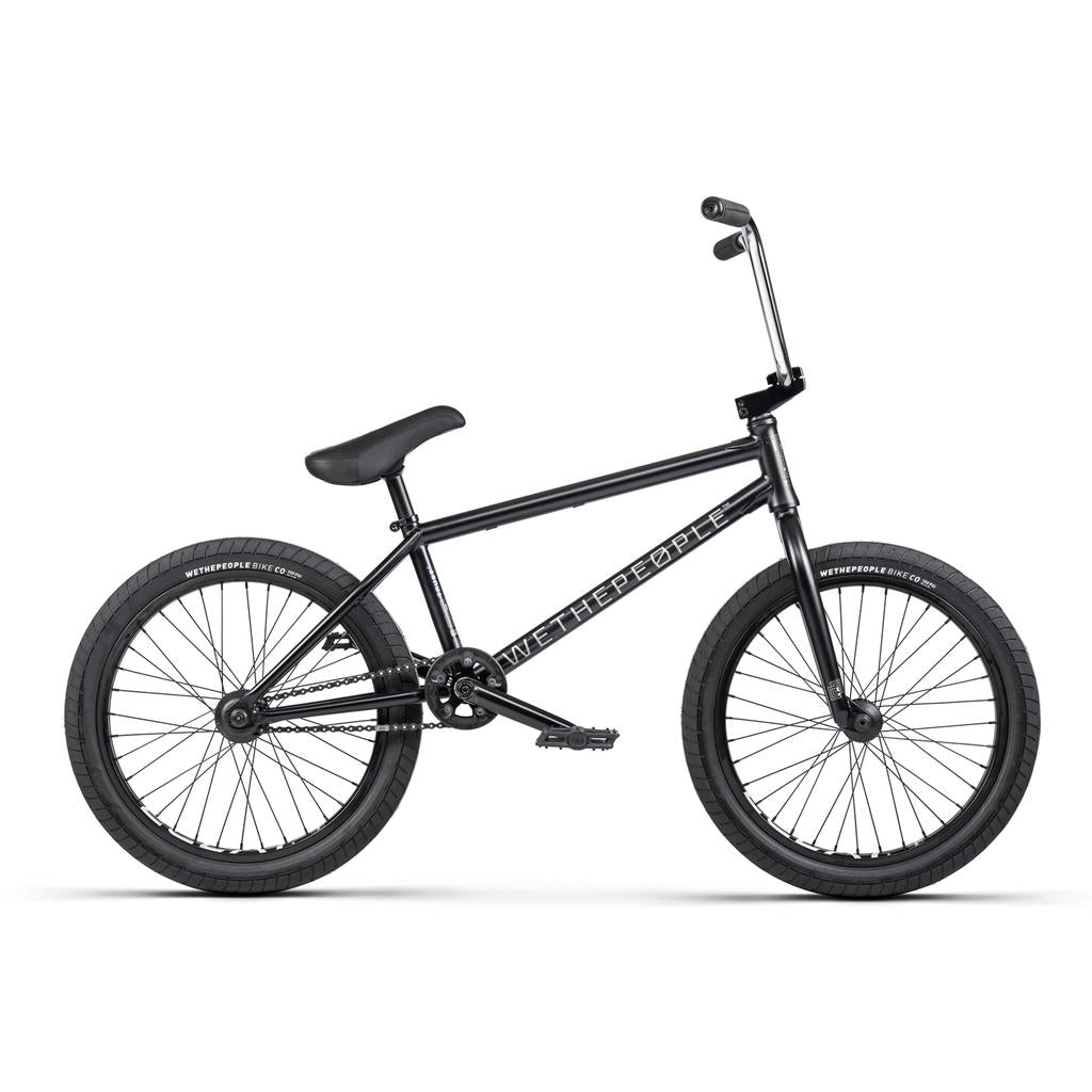 A black Wethepeople Trust 20 Inch Cassette/Hybrid Bike with thick tires, padded seat, straight handlebars, and "We The People" on the frame is shown in side profile against a white background.